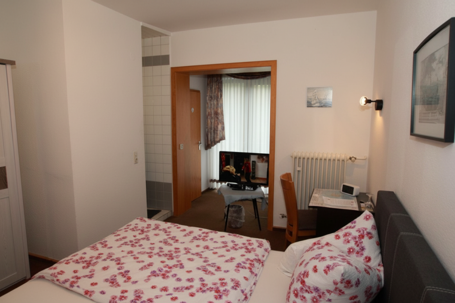 Kleine Hotelzimmer mit Doppelbett, Schreibtisch und Fenster im Hintergrund.